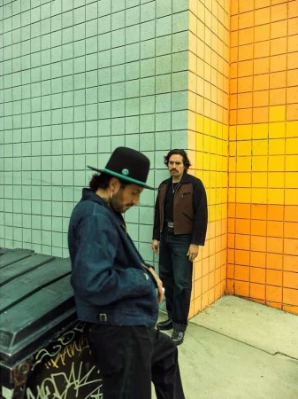 Two men posing outdoors by a colorful tiled wall. One man in a hat leans on a container, while the other stands in the background.