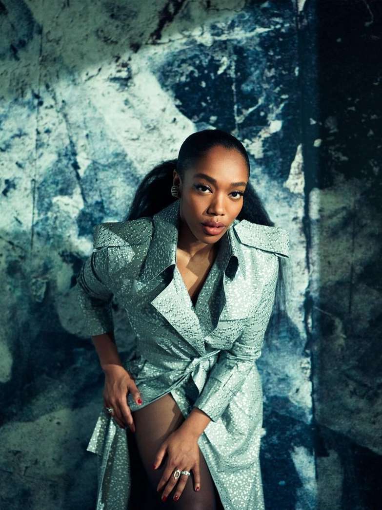Woman in a silver coat posing against a blue and white marbled wall. The lighting and composition highlight her fashion and beauty.