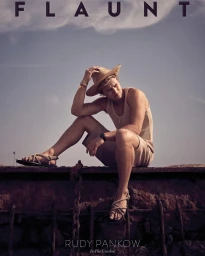 Actor Rudy Pankow wearing a straw hat and beige tank top seated on a metal ledge against a cloudy sky