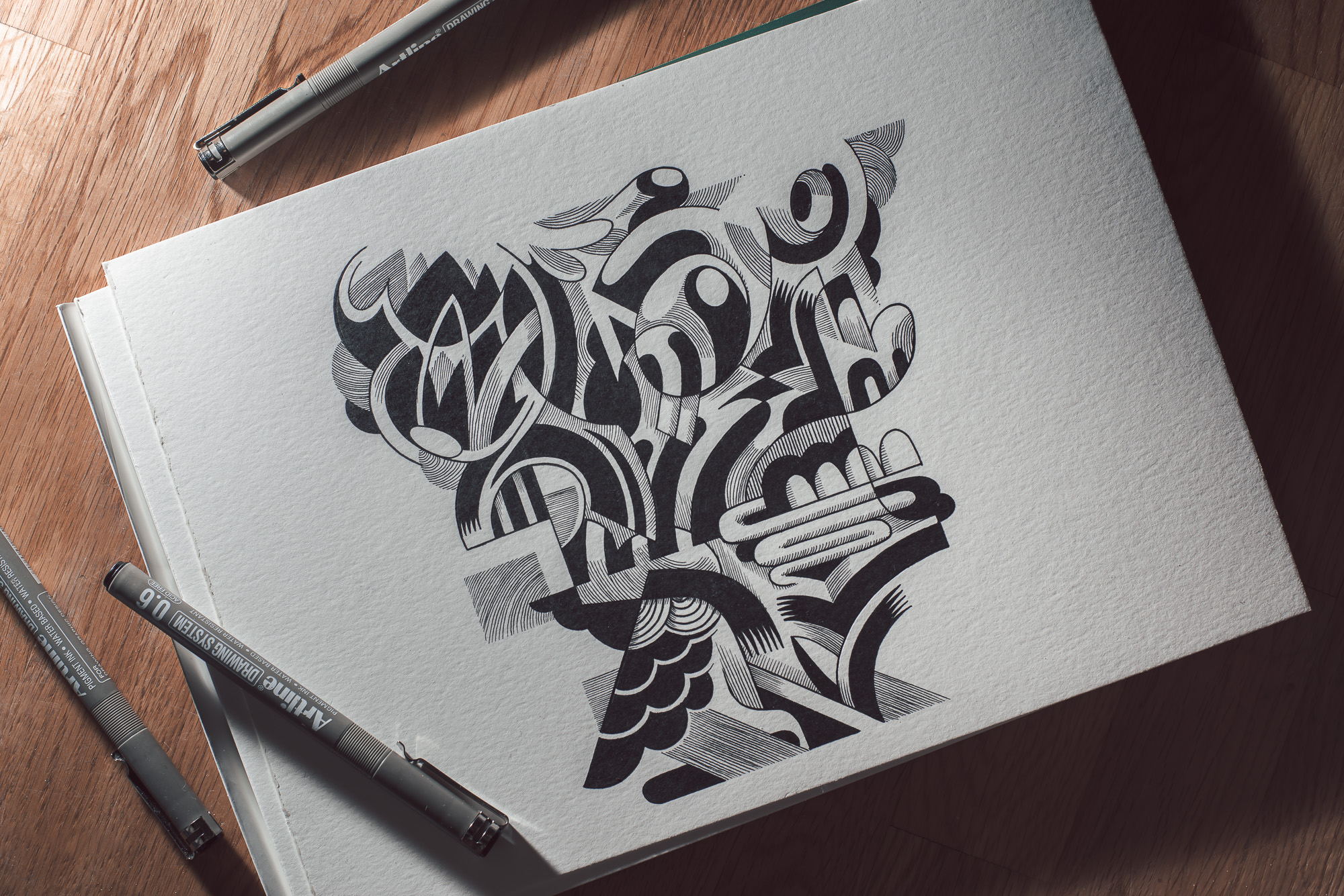 Abstract drawing on white paper featuring black ink pattern; sketch lies on wooden table with grey drawing pens nearby.
