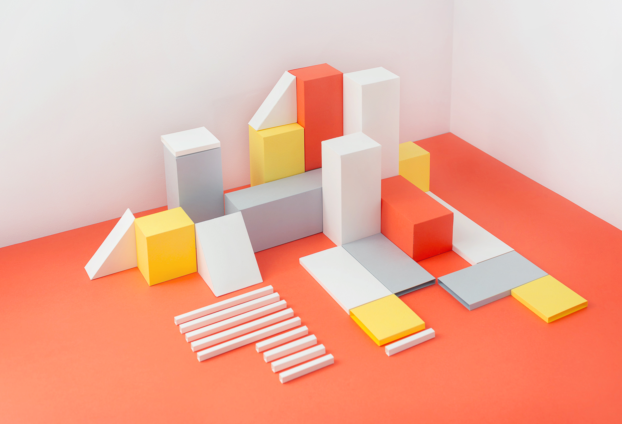 Geometric composition features white, grey, yellow, and red blocks arranged on a coral-colored surface in a well-lit corner.