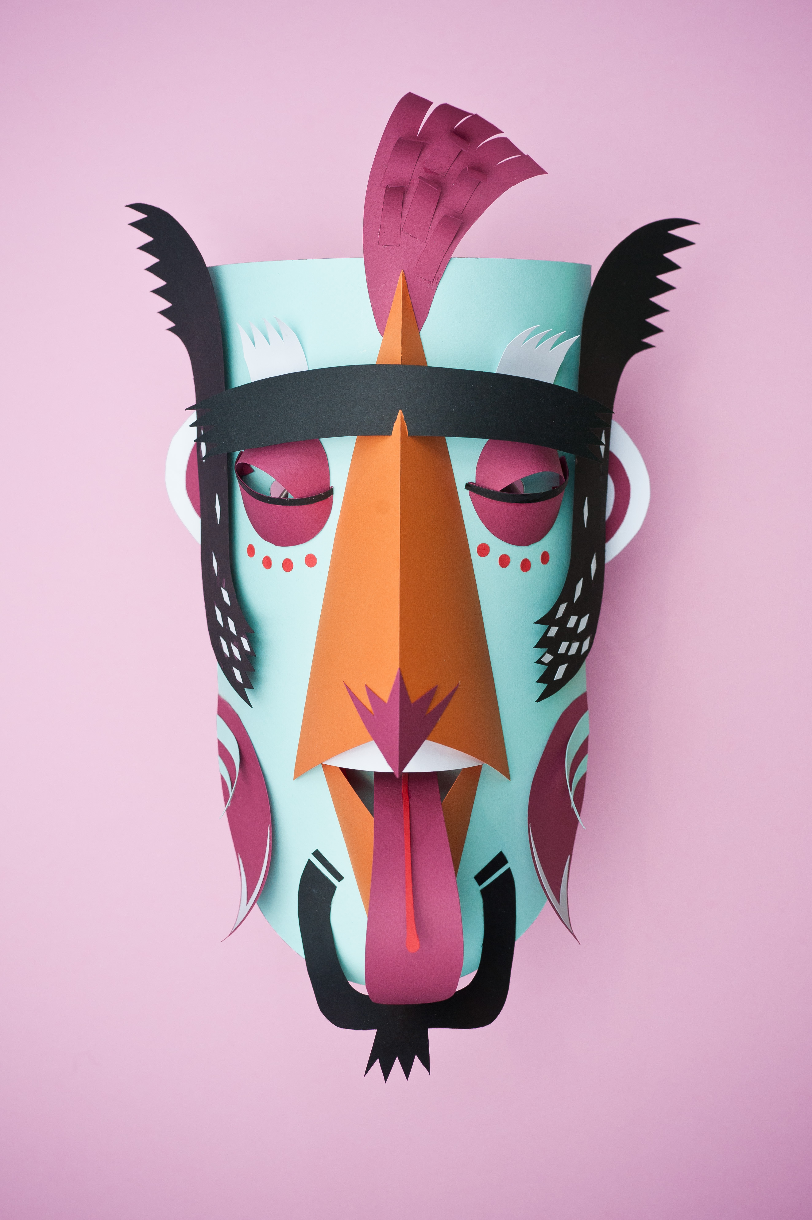 Handmade paper mask art with a light blue base, orange nose, dark pink accents, and a black headdress on a pink background.