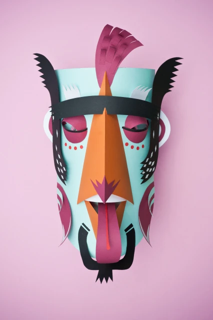 Handmade paper mask art with a light blue base, orange nose, dark pink accents, and a black headdress on a pink background.