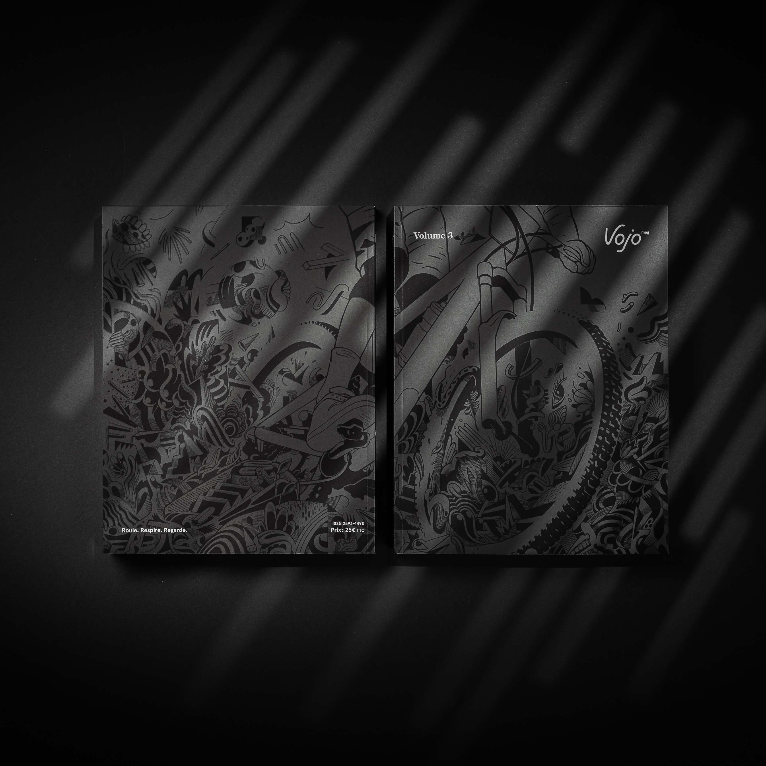 Two copies of VOJO Volume 3 books with black cover art featuring bicycle illustrations on a dark gray surface with striped shadows.
