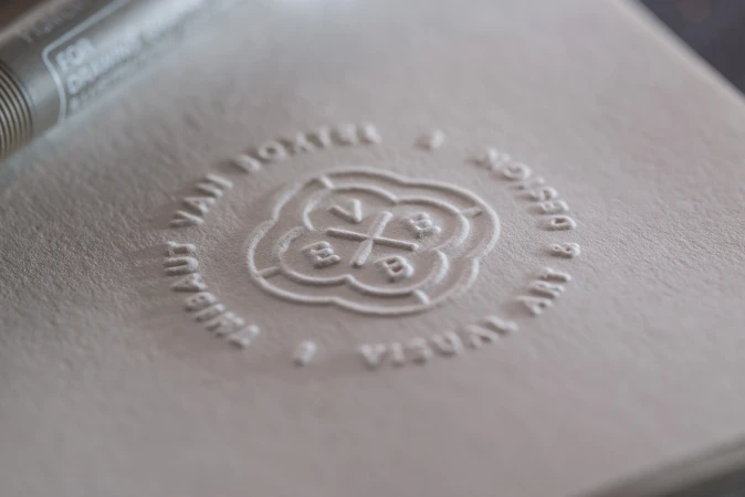 Close-up of an embossed logo on textured paper with visible brand initials and surrounding text, soft lighting, studio environment.