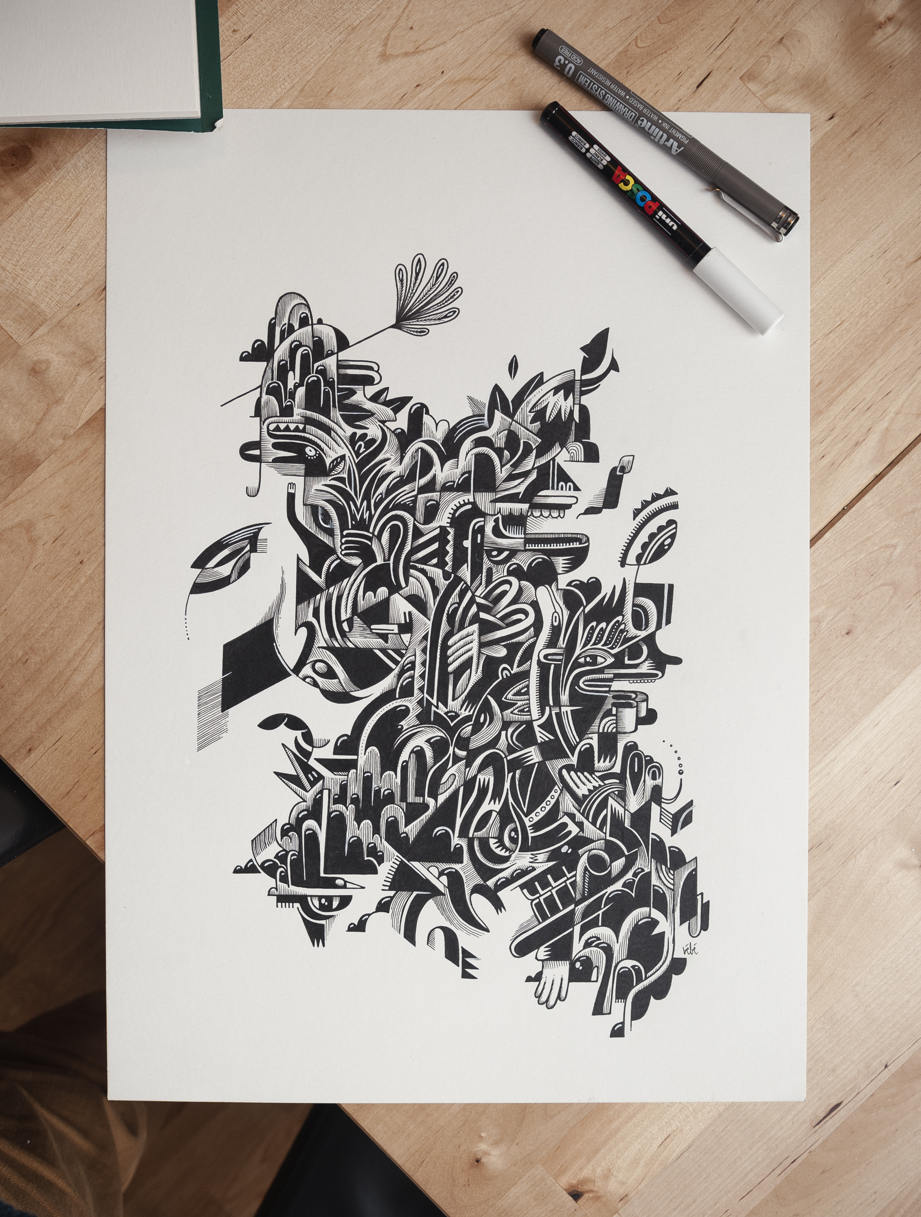 Abstract ink drawing on paper featuring a complex composition of black and white shapes and lines. The artwork has two pens nearby.