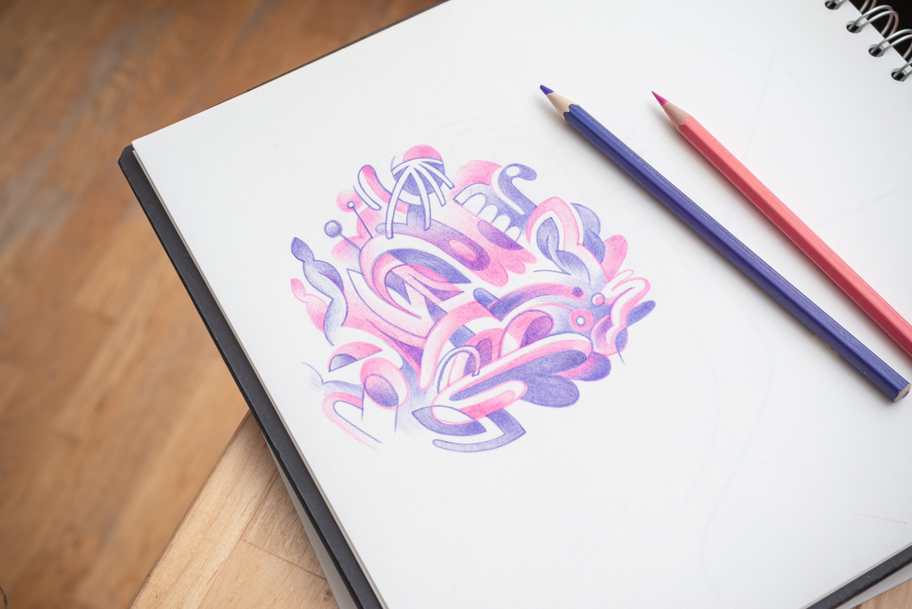 Close-up of a sketchbook with a pink and purple abstract design, and two colored pencils, one purple and one pink, on a wood surface.