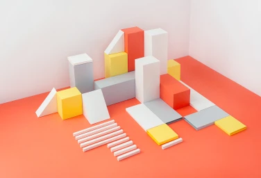 Arrangement of colorful geometric paper blocks, prisms, and cuboids on a coral-orange surface against white walls.