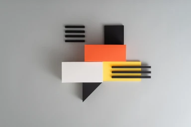 Three-dimensional geometric wall composition of orange, yellow, white, and black rectangular blocks and bars against a grey wall.