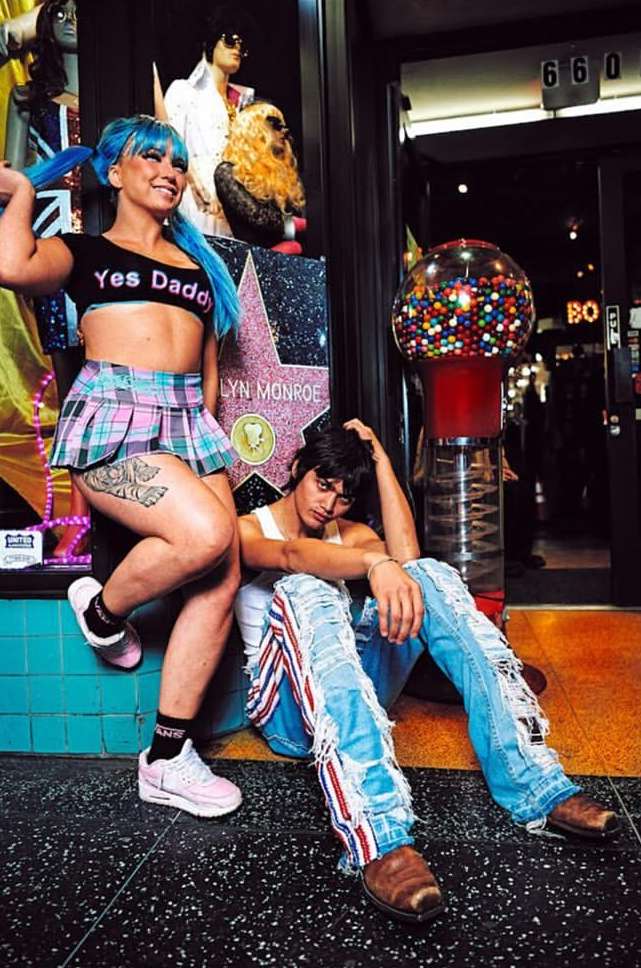A woman with blue pigtails and a man in ripped jeans pose next to a Hollywood Walk of Fame star and a gumball machine at night.
