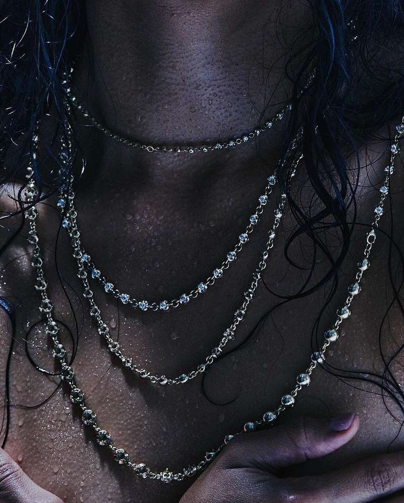 Close-up of a person's chest and neck, adorned with multiple beaded necklaces, water droplets on skin and dark hair visible.