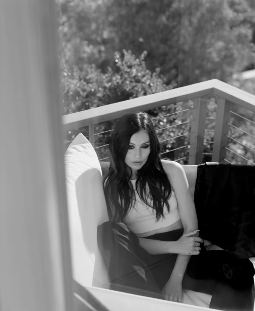 Monochrome portrait of a woman with long dark hair, wearing a crop top and pants, sitting on a white couch on an outdoor deck.