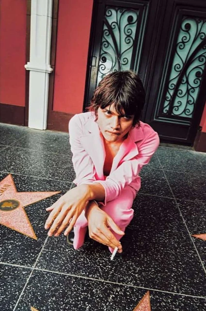 Adult male in a pink suit kneeling on a star-lined sidewalk holds a cigarette in an urban setting, looking directly at the camera.