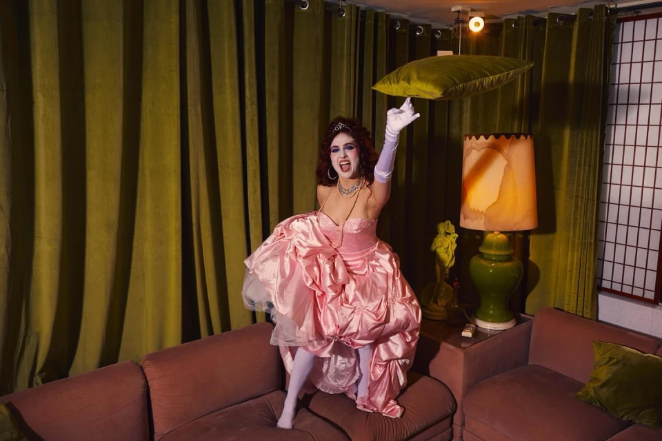 Woman in ruffled pink gown poses on maroon couch with green velvet pillow under a hanging green velvet pillow and vintage lamp indoors.