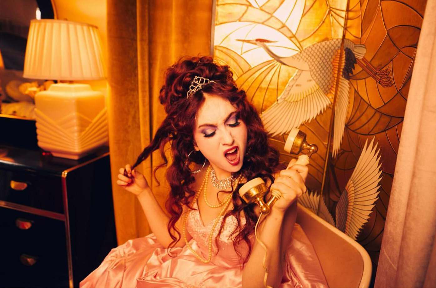 Glamorous woman in pink dress yelling into vintage telephone, styled with tiara, jewelry, and updo, set against ornate backdrop.