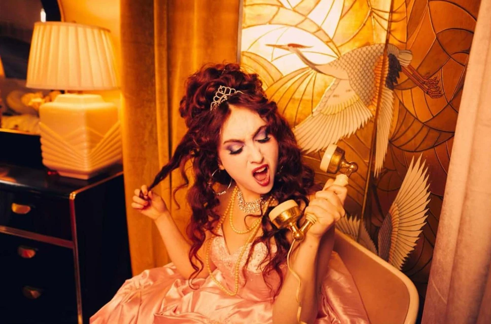 Glamorous woman in pink dress yelling into vintage telephone, styled with tiara, jewelry, and updo, set against ornate backdrop.