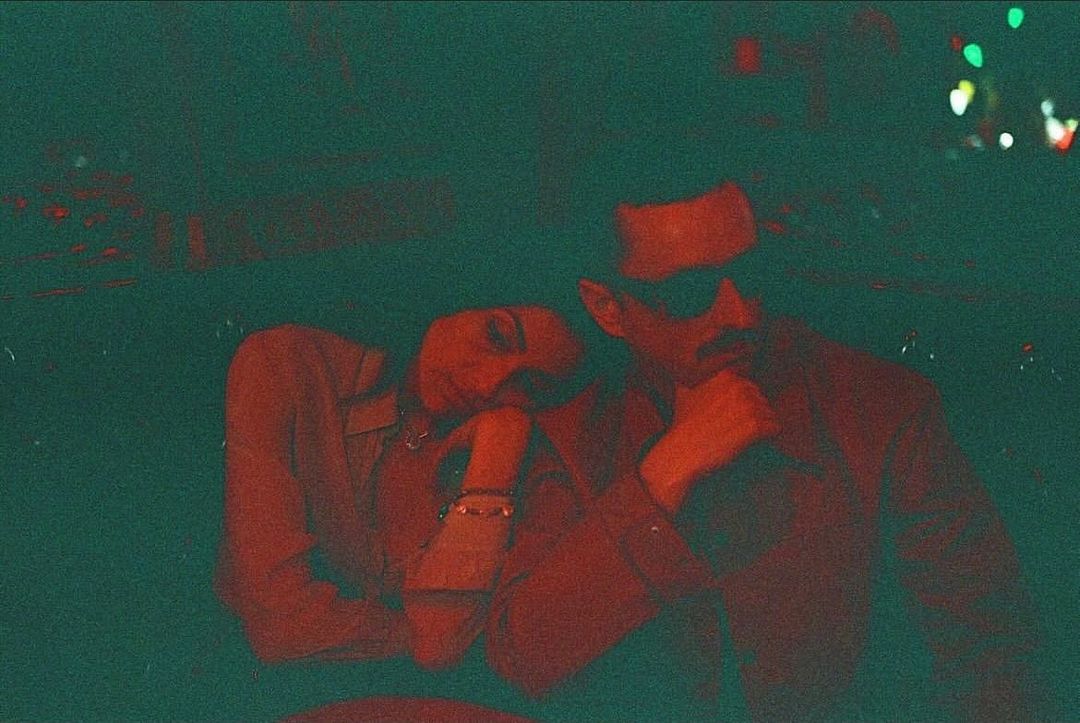 A man and a woman are posing indoors in a low-light photo, showcasing a dark, moody aesthetic with the color red dominating the image.