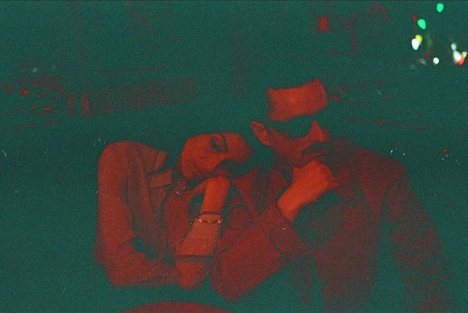 A man and a woman are posing indoors in a low-light photo, showcasing a dark, moody aesthetic with the color red dominating the image.