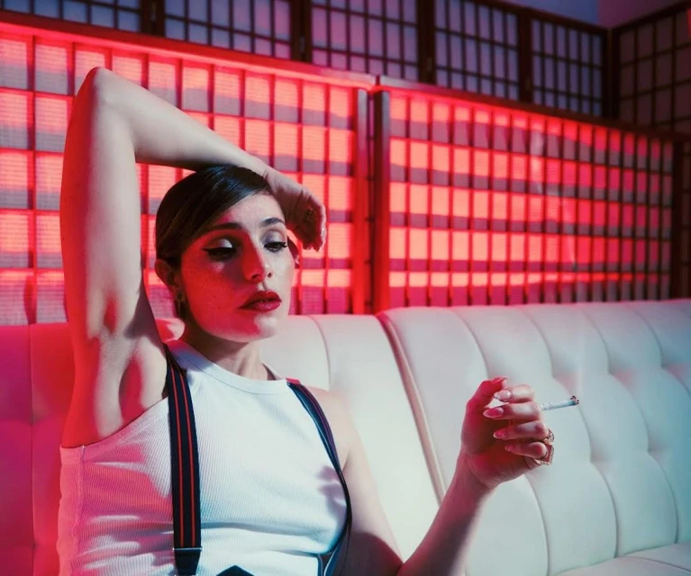 A woman with dark hair and red lipstick reclines on a white couch, holding a cigarette against a red-lit geometric background, in a stylish indoor setting.