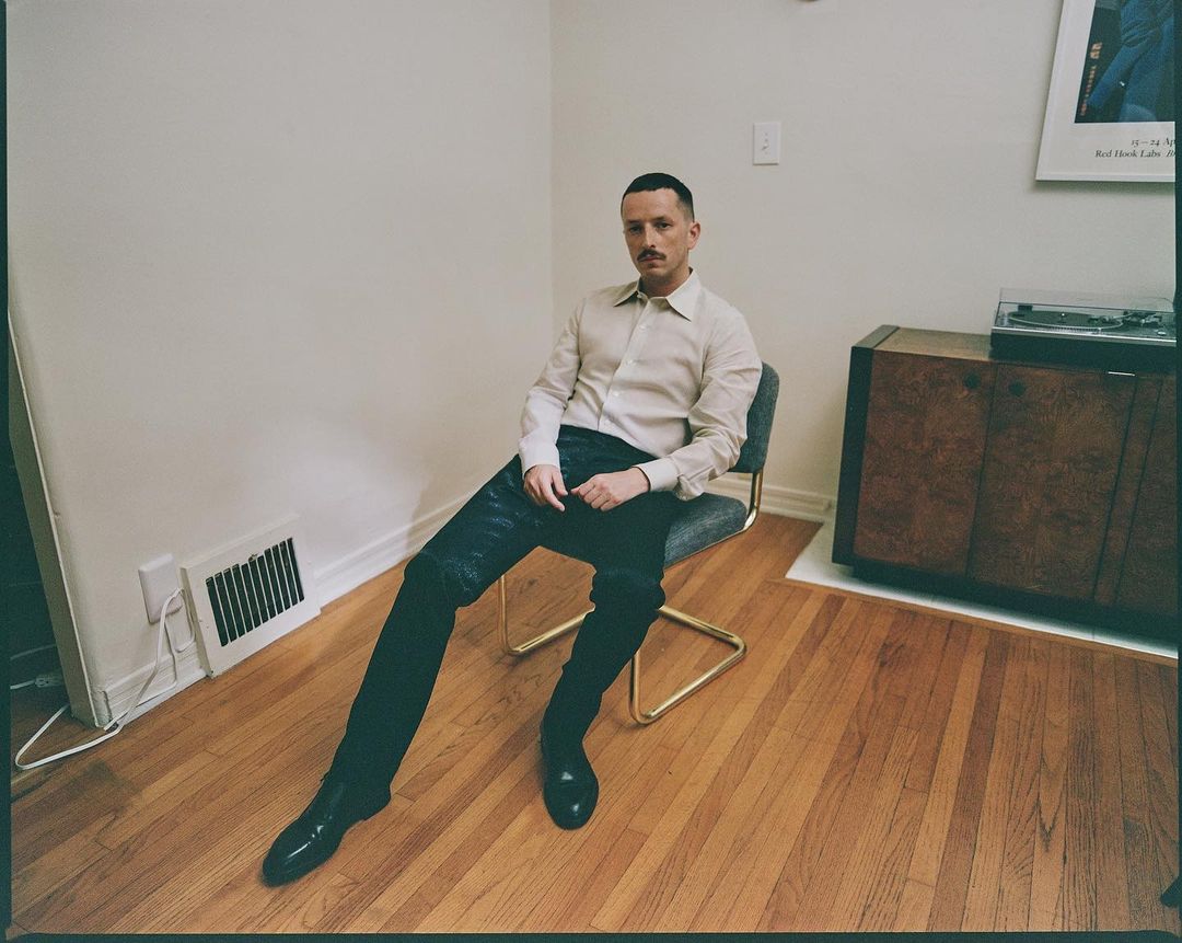 Stylish man in a dress shirt and shiny black pants sitting on a chair in a minimalist interior with hardwood floors and white walls.
