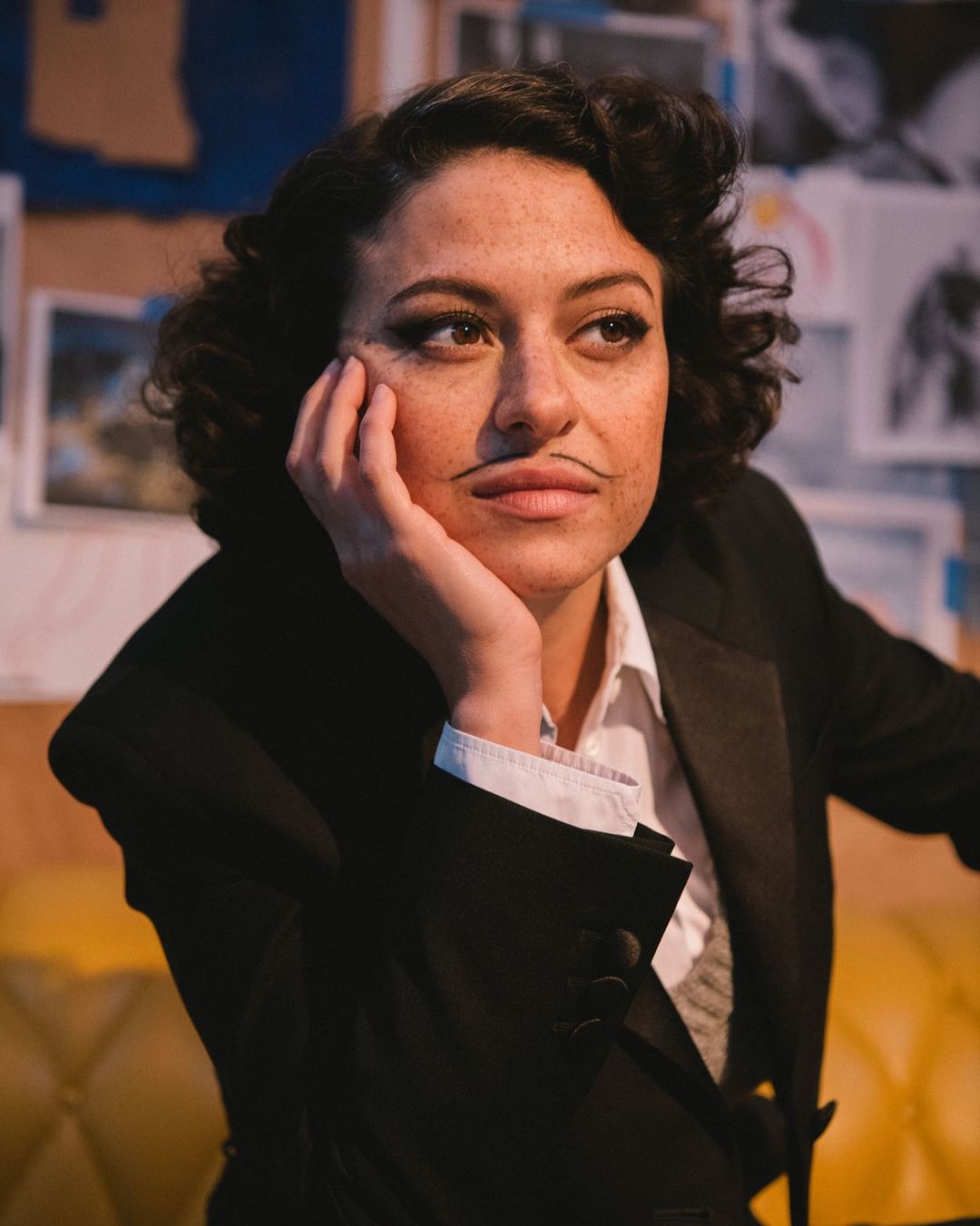 Woman with drawn-on mustache in studio. Portrait of a person with curly dark hair wearing a black jacket and white shirt.