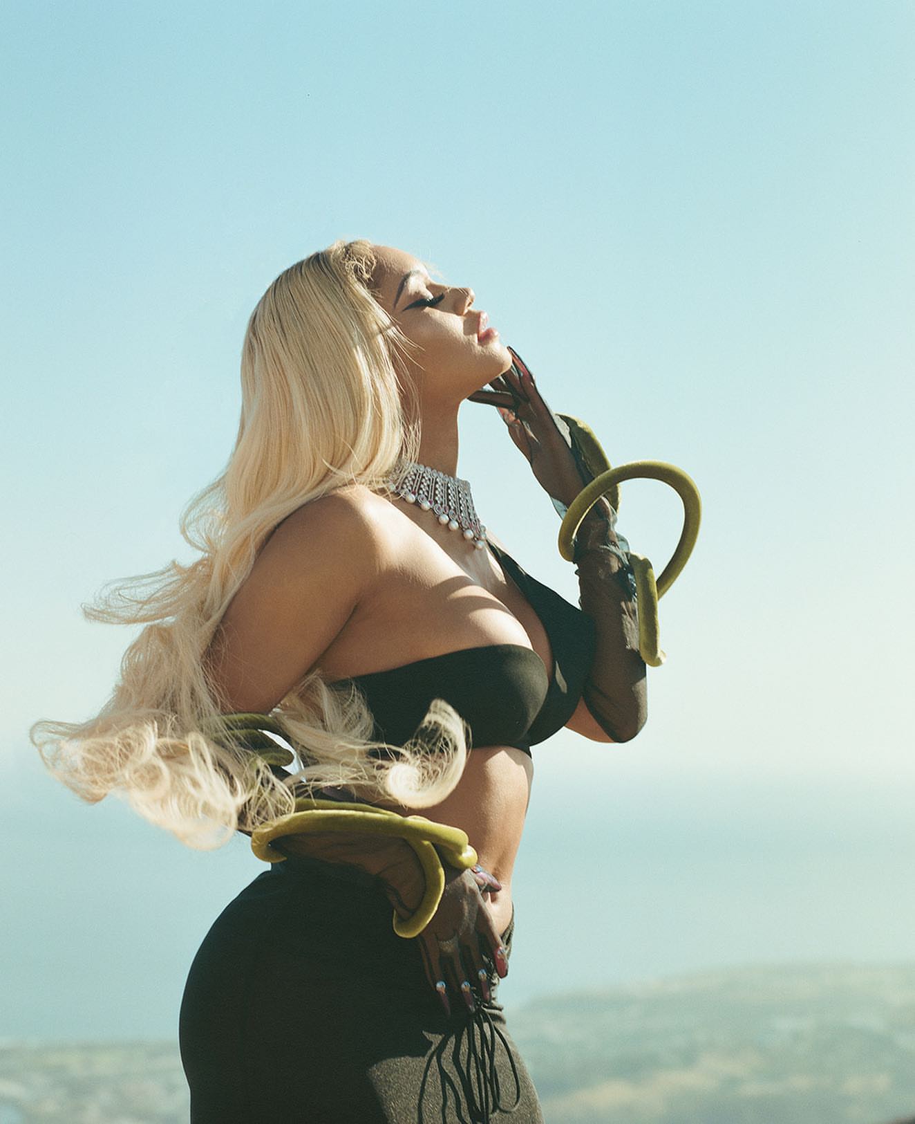 An adult female poses outdoors in a black form-fitting dress with blond wavy hair. She wears jewelry with olive arm bands against a sky backdrop.