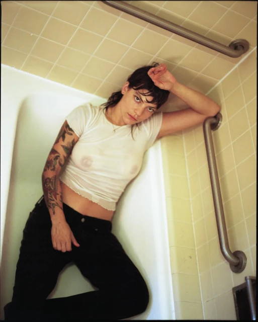 Woman with tattoos lounges in a white tiled bathtub, wearing a white t-shirt and black pants, under soft lighting.
