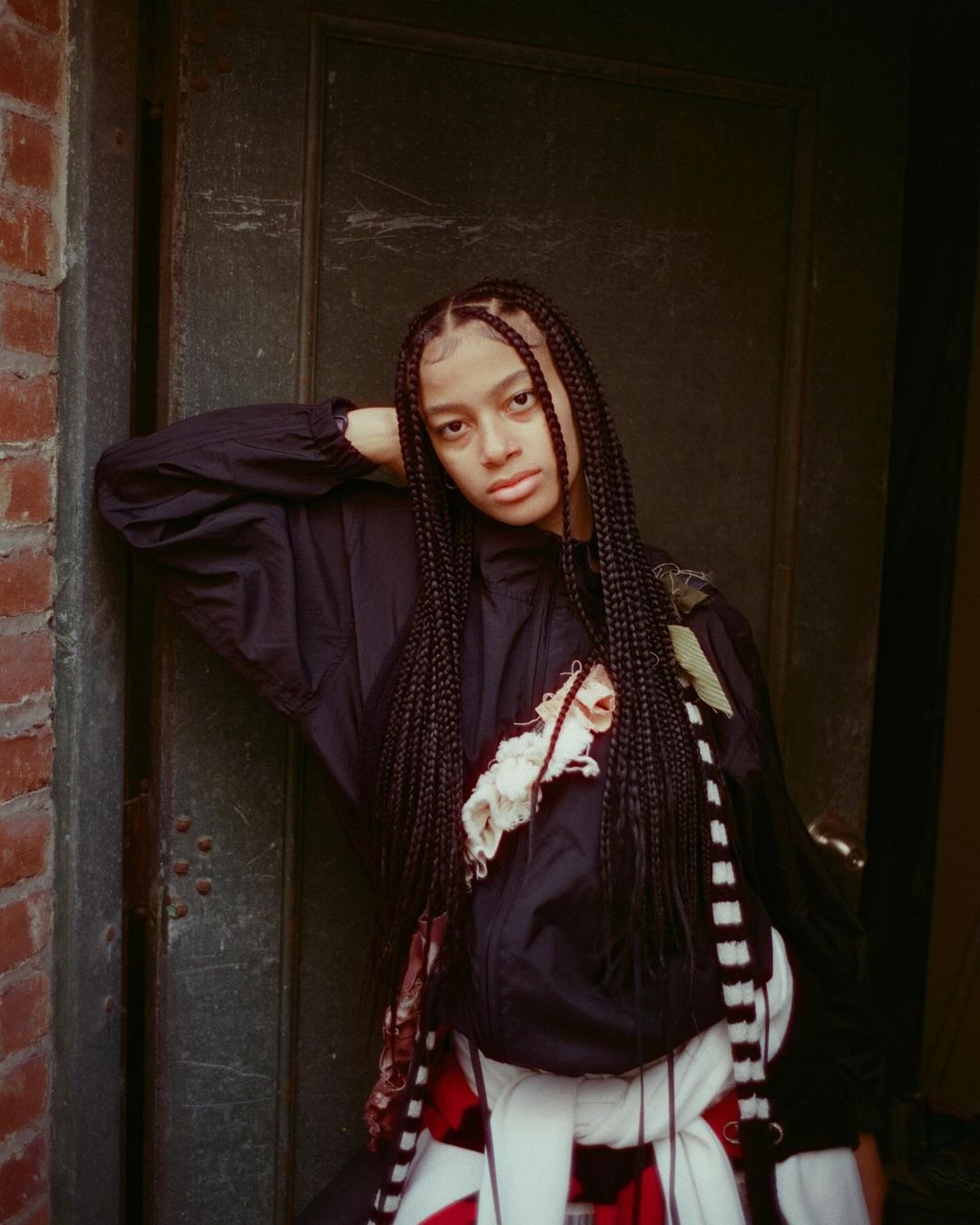 Young woman with braids poses next to a dark green door on a brick building wearing a black jacket and a red and white scarf.