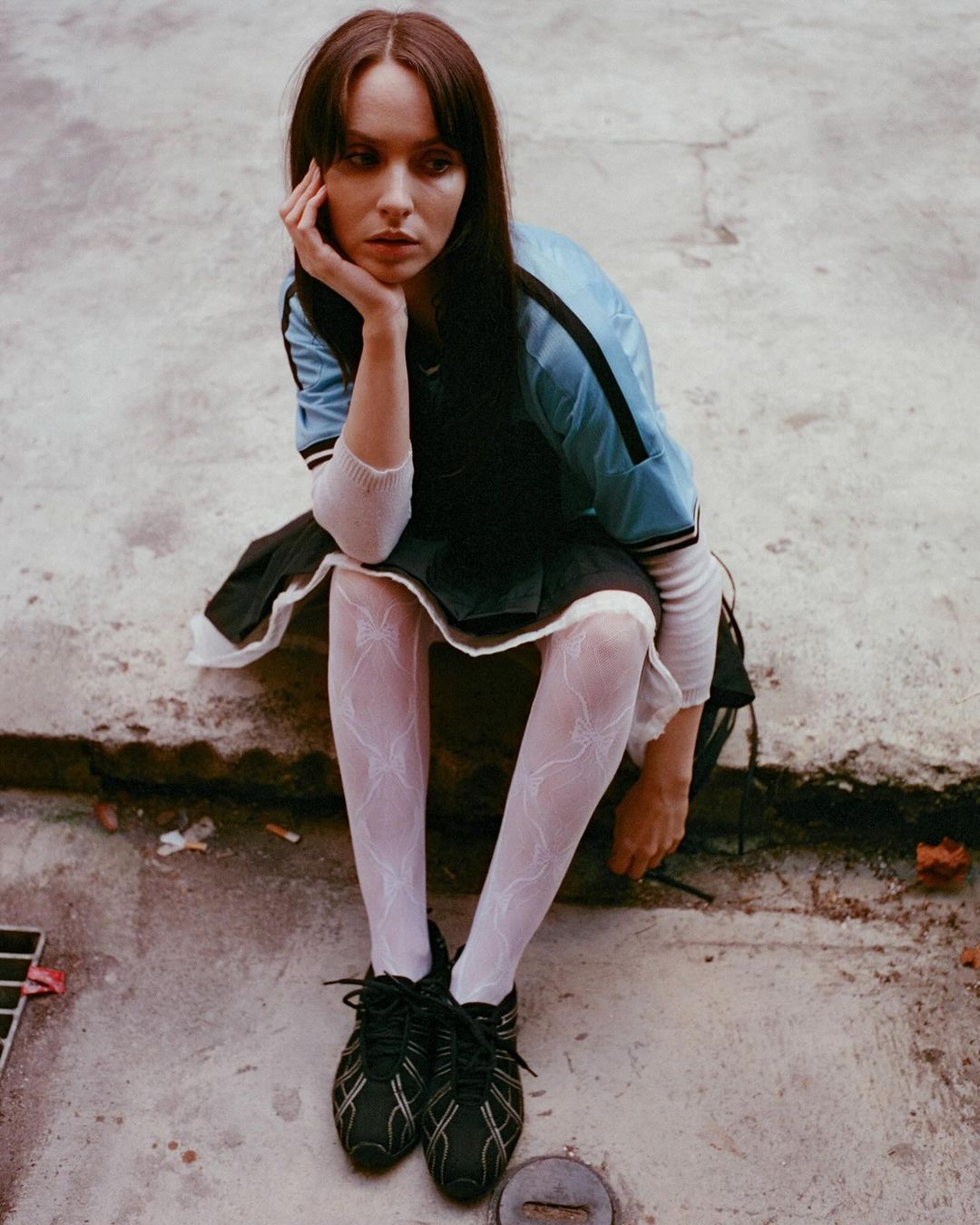 A young woman with long dark hair sits on concrete in a blue jacket, black skirt, patterned tights, and black shoes in natural light.
