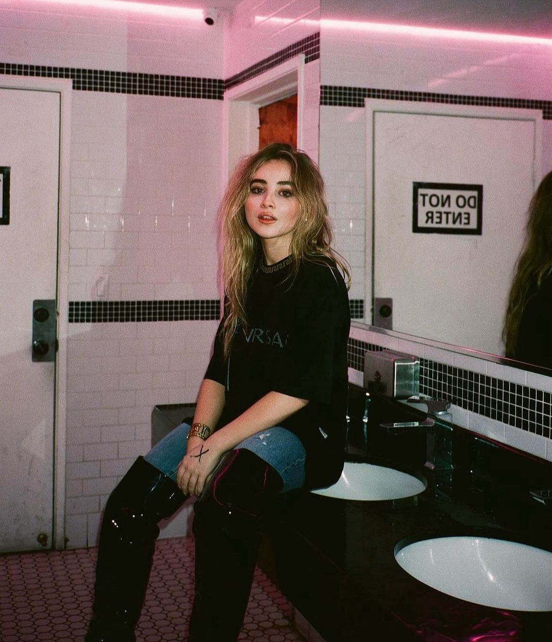 Adult female posing in a bathroom with black and white tile, illuminated by pink neon lighting, wearing black shirt and high black boots.