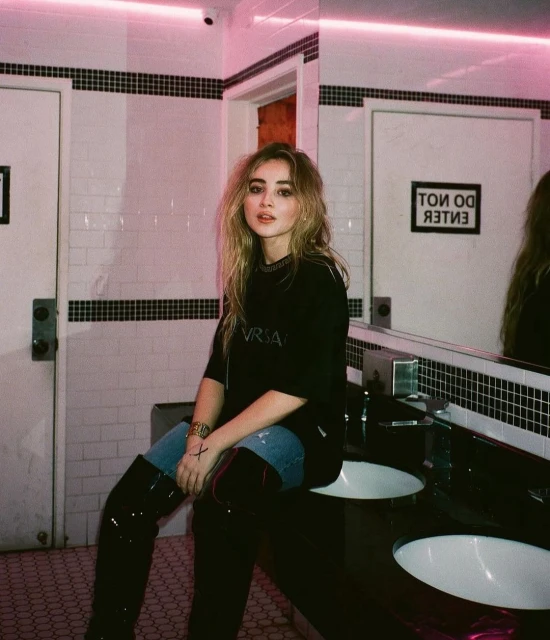 Adult female posing in a bathroom with black and white tile, illuminated by pink neon lighting, wearing black shirt and high black boots.