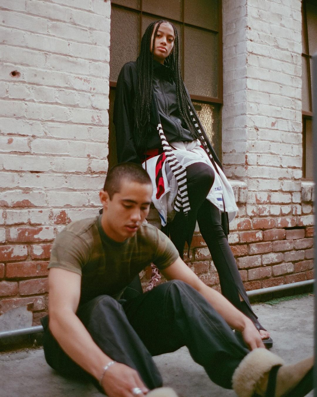 Two people pose outdoors against a brick wall; an adult male sits on the ground, and an adult female leans against the wall, both wearing stylish clothing.