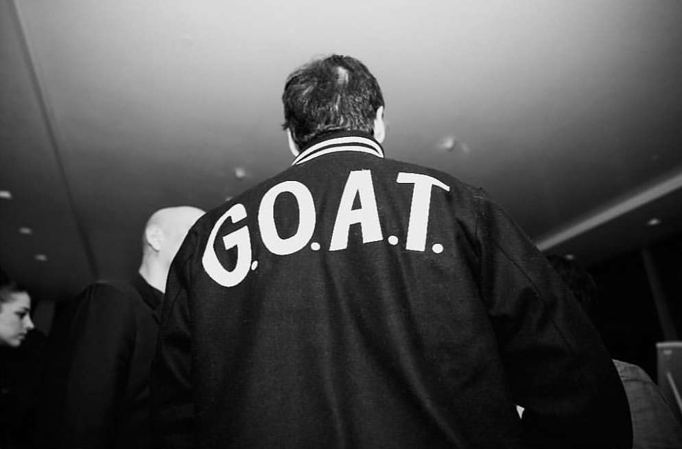 Black and white shot of a group, one adult male wearing a black jacket with 'G.O.A.T.' printed in white across the back.