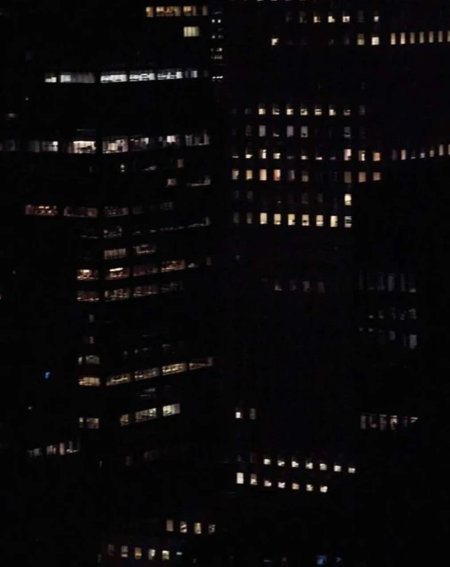 Nighttime view of multiple dark skyscrapers with illuminated windows, creating a grid-like pattern of light in an urban environment.