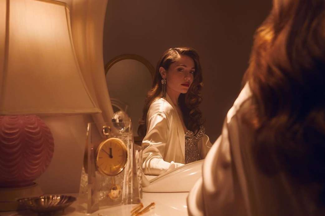 Adult woman with long brown hair reflected in mirror next to a table lamp and a crystal clock in an indoor setting.