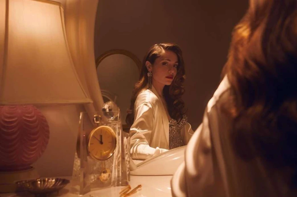 Adult woman with long brown hair reflected in mirror next to a table lamp and a crystal clock in an indoor setting.