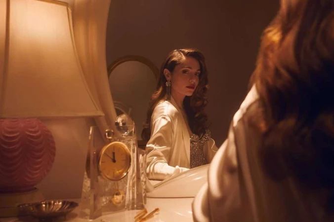 Adult woman with long brown hair reflected in mirror next to a table lamp and a crystal clock in an indoor setting.