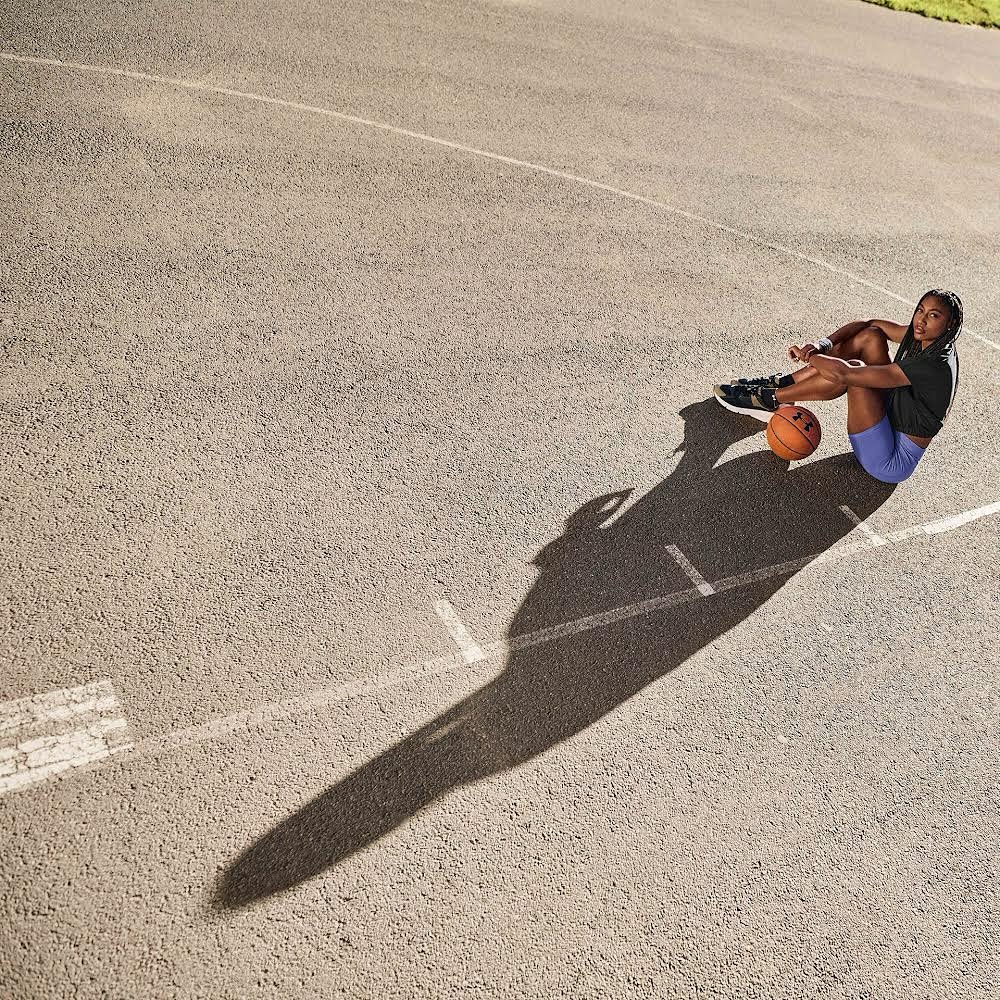 Female athlete sits on outdoor basketball court, resting with basketball. Overhead shot captures full body, surface details, athletic wear.