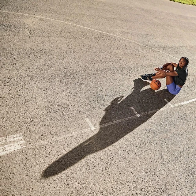 Female athlete sits on outdoor basketball court, resting with basketball. Overhead shot captures full body, surface details, athletic wear.