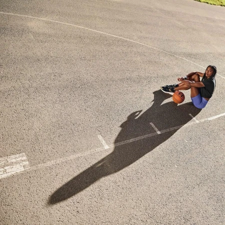 Female athlete sits on outdoor basketball court, resting with basketball. Overhead shot captures full body, surface details, athletic wear.