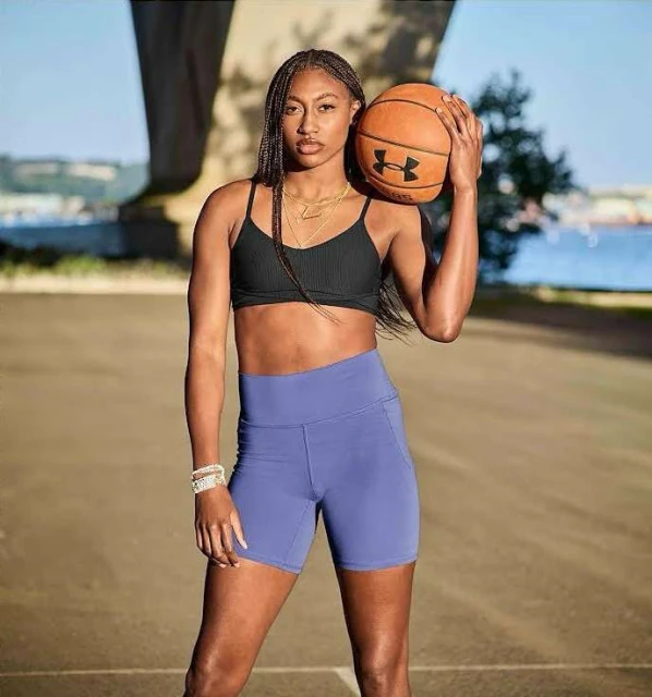 Athletic woman in activewear poses outdoors with a basketball. She wears a sports bra and blue bike shorts with urban background.