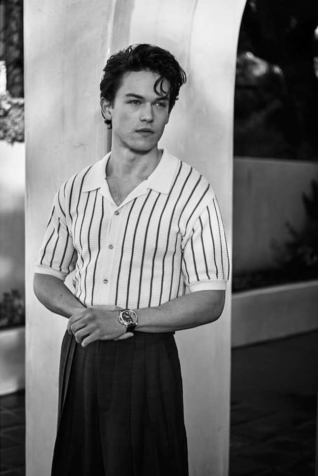 Black and white portrait of a young adult male in a vertically striped collared shirt and dark skirt, standing against an archway.