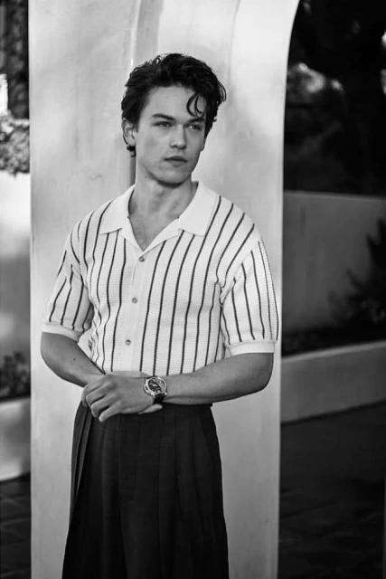 Black and white portrait of a young adult male in a vertically striped collared shirt and dark skirt, standing against an archway.