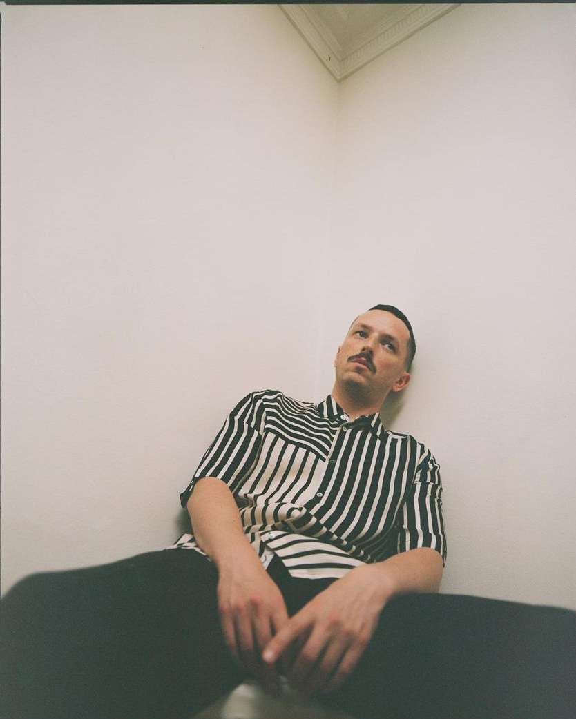 Man in striped shirt sitting against white walls. Indoor fashion portrait of a male model with short dark hair and a mustache.
