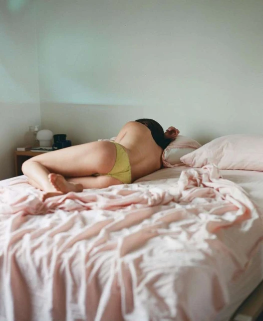 Adult female lies on a pink bed in an indoor setting. The woman has dark hair and wears yellow underwear, facing away from the camera.