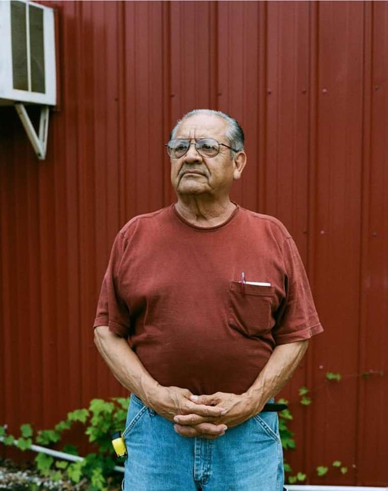 Portrait of an elderly man with glasses in front of a red metal wall, wearing a rust-colored t-shirt and blue jeans in an outdoor setting.
