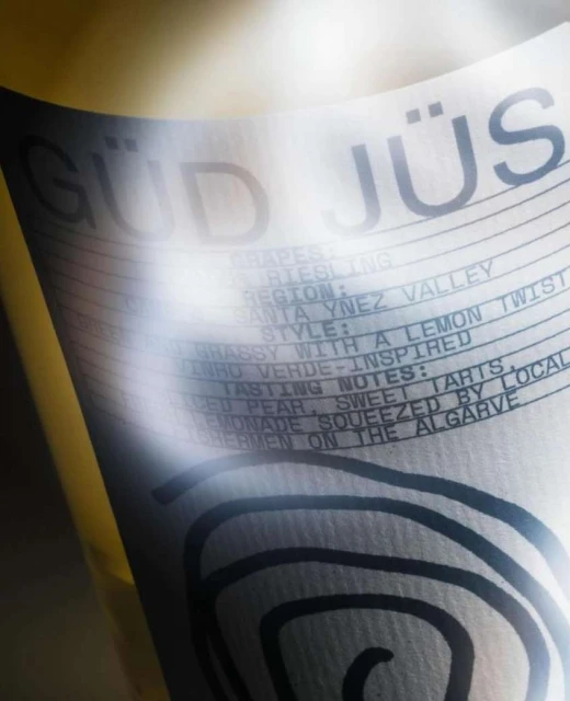 Close-up of a wine bottle label with the name 'GÜD JÜS' and tasting notes like 'lemon twist' and 'squeezed by local fishermen'.