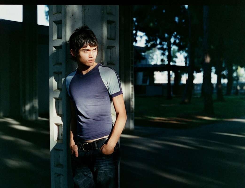 Young man in a blue t-shirt stands near a concrete pillar in partial sunlight, looking off to the side, with a park in the background.