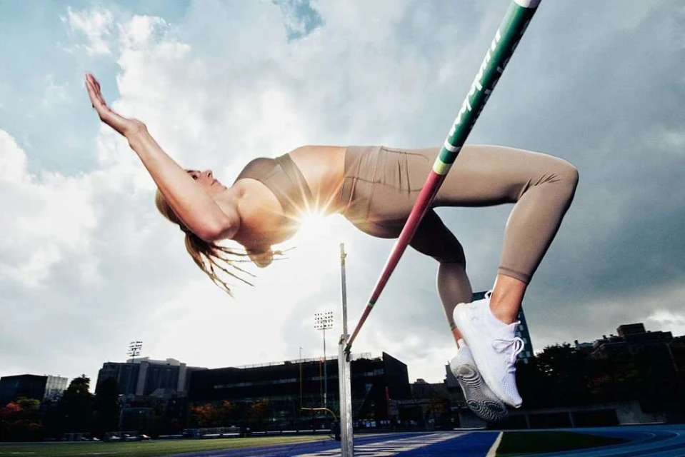 Female athlete in mid-air executing the high jump over a horizontal bar, with stadium seating and cloudy sky in the background.