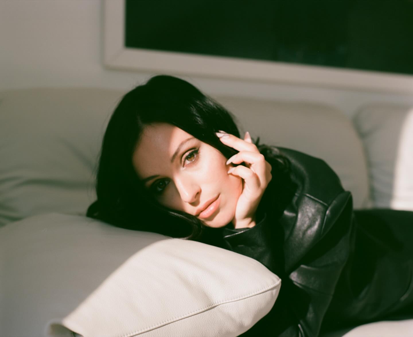 Portrait of a woman with long dark hair and a black jacket lying on a cream-colored couch indoors, with sunlight streaming in.